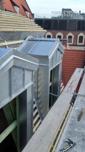 Montaż metalowych obróbek blacharskich kominów na dachu budynku wielorodzinnego, widoczne rusztowania i narzędzia pracy.