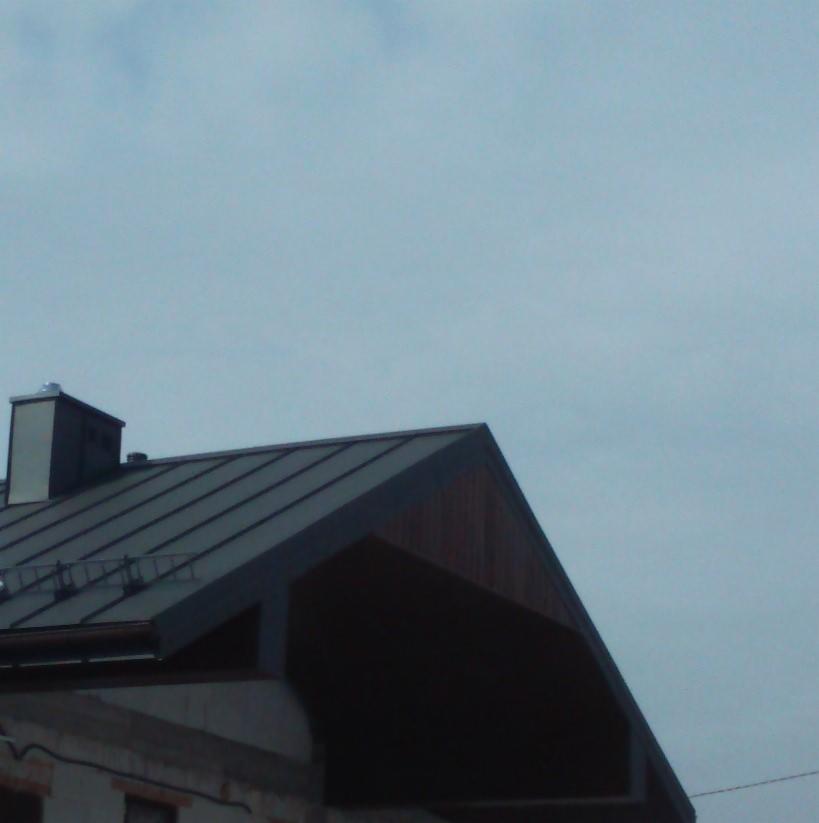 Fragment dachu z blachy na budynku w trakcie budowy, widoczny komin i drewniane wykończenie szczytu dachu na tle pochmurnego nieba.