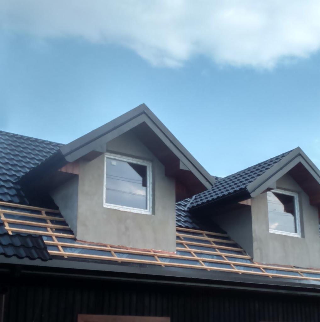 Montaż dachu z czarnej blachodachówki na domu z oknami dachowymi, widoczne drewniane łaty i folia dachowa w trakcie prac.