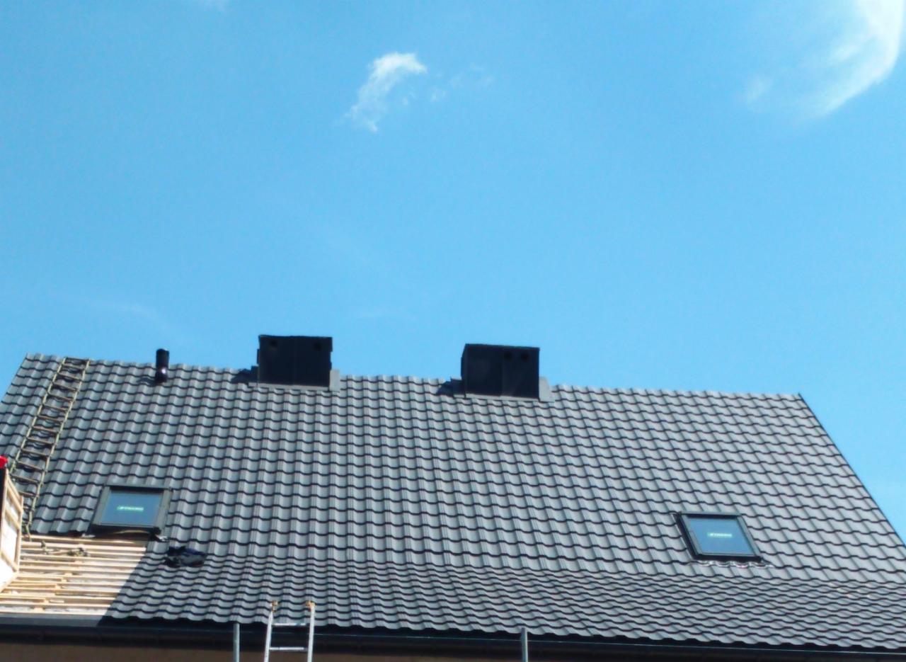 Szary dach z dachówki ceramicznej z dwoma kominami i oknami dachowymi, fragment rusztowania i drabina oparta o budynek.