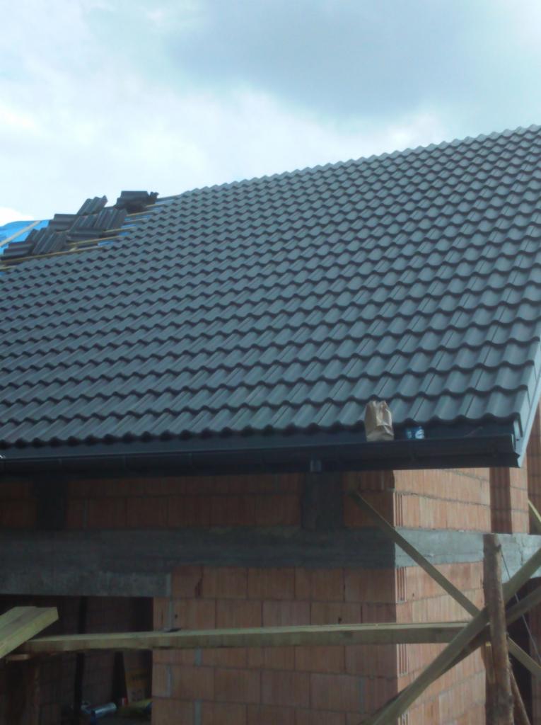 Częściowo pokryty dach z ciemnej dachówki na surowym budynku z czerwonej cegły, widoczne rusztowanie i resztki materiałów budowlanych na dachu.