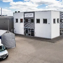 Budynek firmy Wenteco, centrum dystrybucji wentylacji, z zaparkowanym samochodem dostawczym Solar Plus, specjalizującym się w wentylacji i klimatyzacji, na tle błękitnego nieba z chmurami.