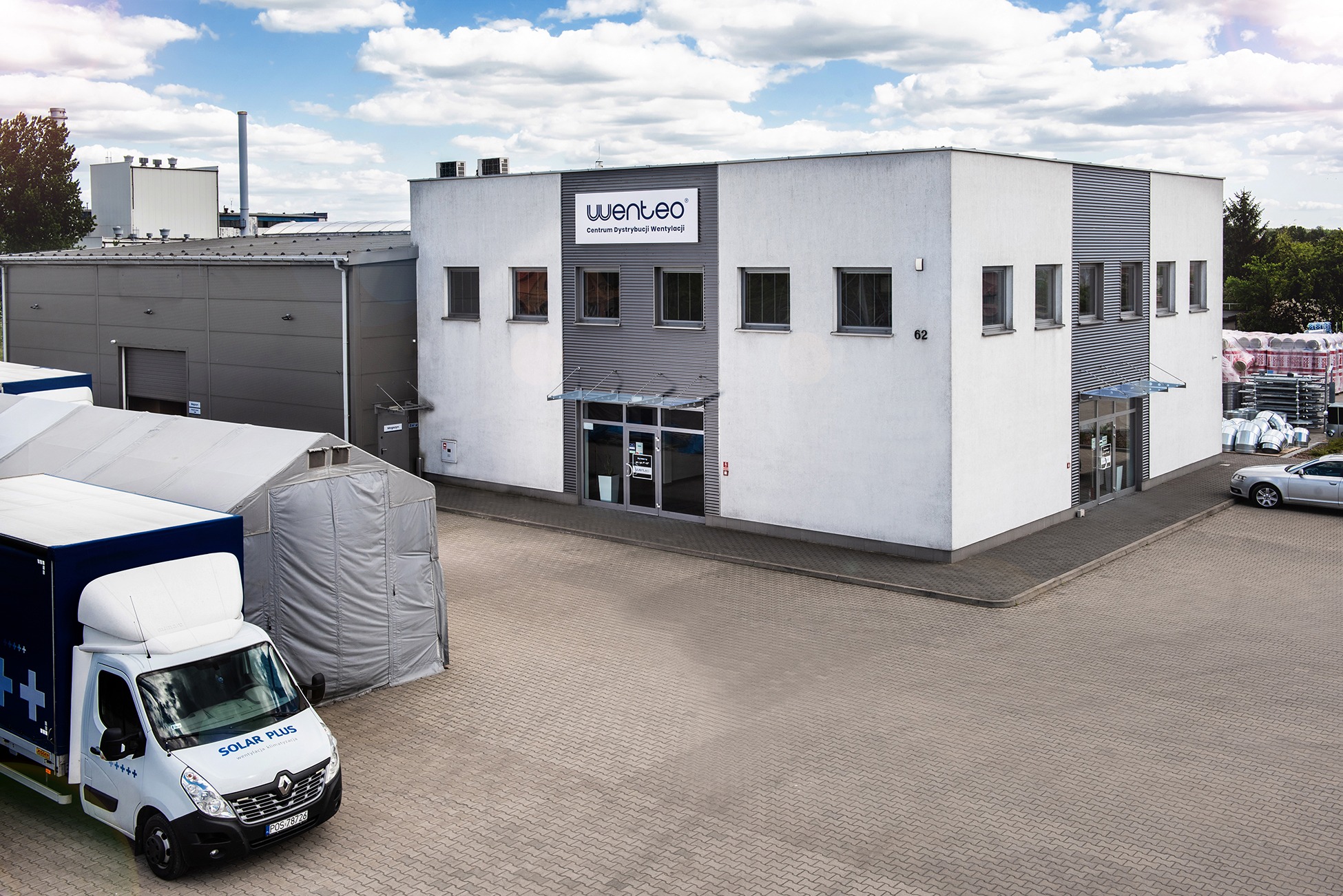 Budynek firmy Wenteco, centrum dystrybucji wentylacji, z zaparkowanym samochodem dostawczym Solar Plus, specjalizującym się w wentylacji i klimatyzacji, na tle błękitnego nieba z chmurami.