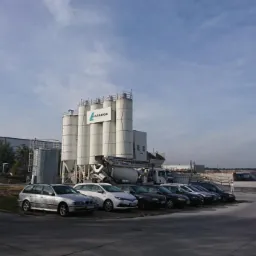 Betoniarnia Lafarge z rzędem zaparkowanych samochodów osobowych na pierwszym planie, widok z poziomu parkingu w słoneczny dzień.