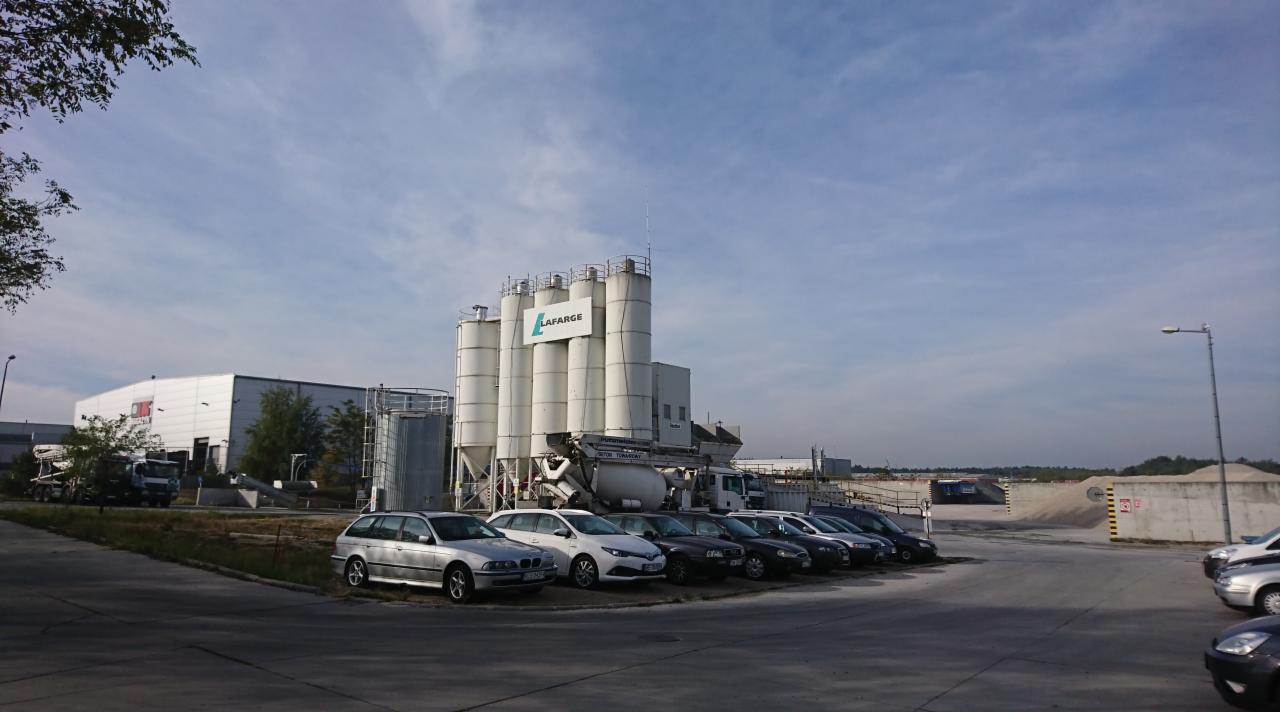 Betoniarnia Lafarge z rzędem zaparkowanych samochodów osobowych na pierwszym planie, widok z poziomu parkingu w słoneczny dzień.