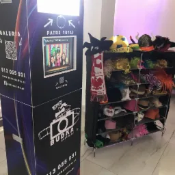 Automat do zdjęć typu budka selfie z ekranem, obok regał z perukami, kapeluszami i rekwizytami do zdjęć, w tle osoby na uroczystości.