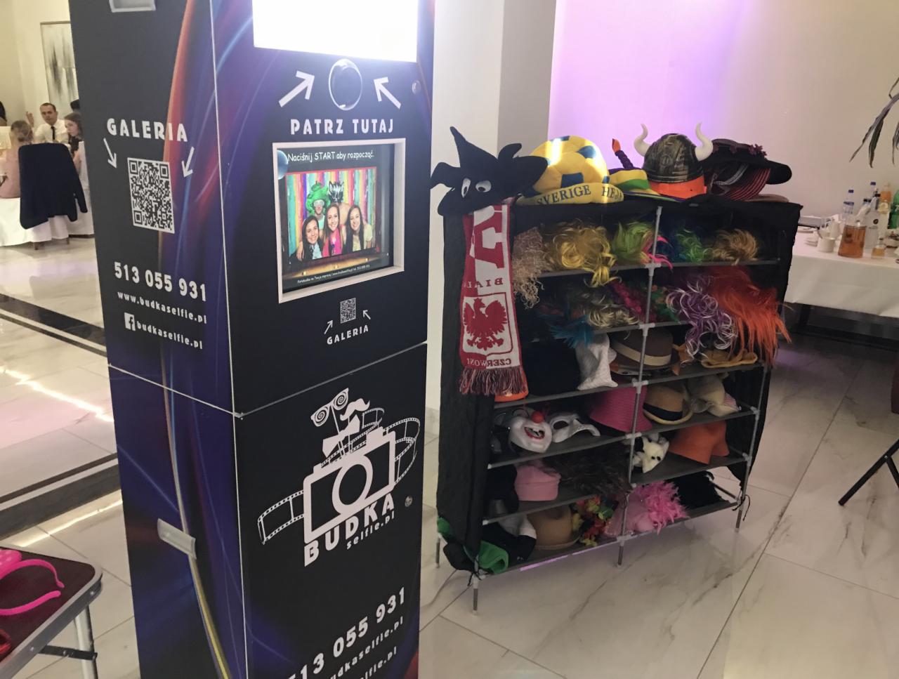 Automat do zdjęć typu budka selfie z ekranem, obok regał z perukami, kapeluszami i rekwizytami do zdjęć, w tle osoby na uroczystości.