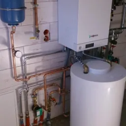Nowoczesny piec gazowy Viessmann z zasobnikiem ciepłej wody, instalacja z rur miedzianych i stalowych, manometr, osprzęt, białe płytki ścienne imitujące drewno.