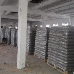 Magazyn pelletu: rzędy zapakowanych palet z pelletem w folii stretch, widoczny pies w tle, betonowe filary i okna magazynu.