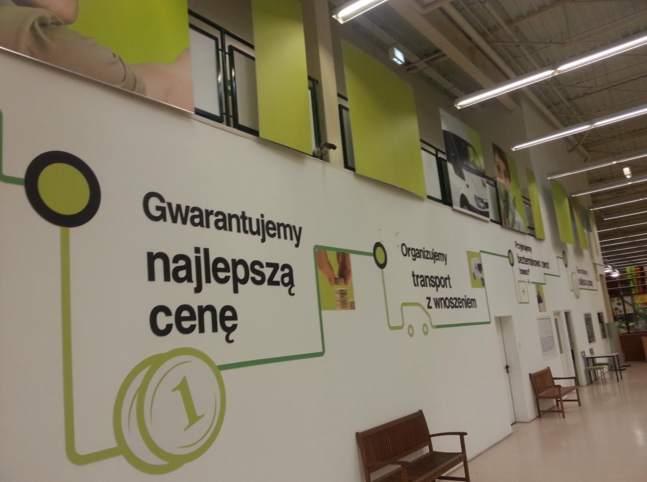 Wnętrze sklepu z grafiką ścienną promującą niskie ceny i transport z wniesieniem, z widocznymi ławkami dla klientów.
