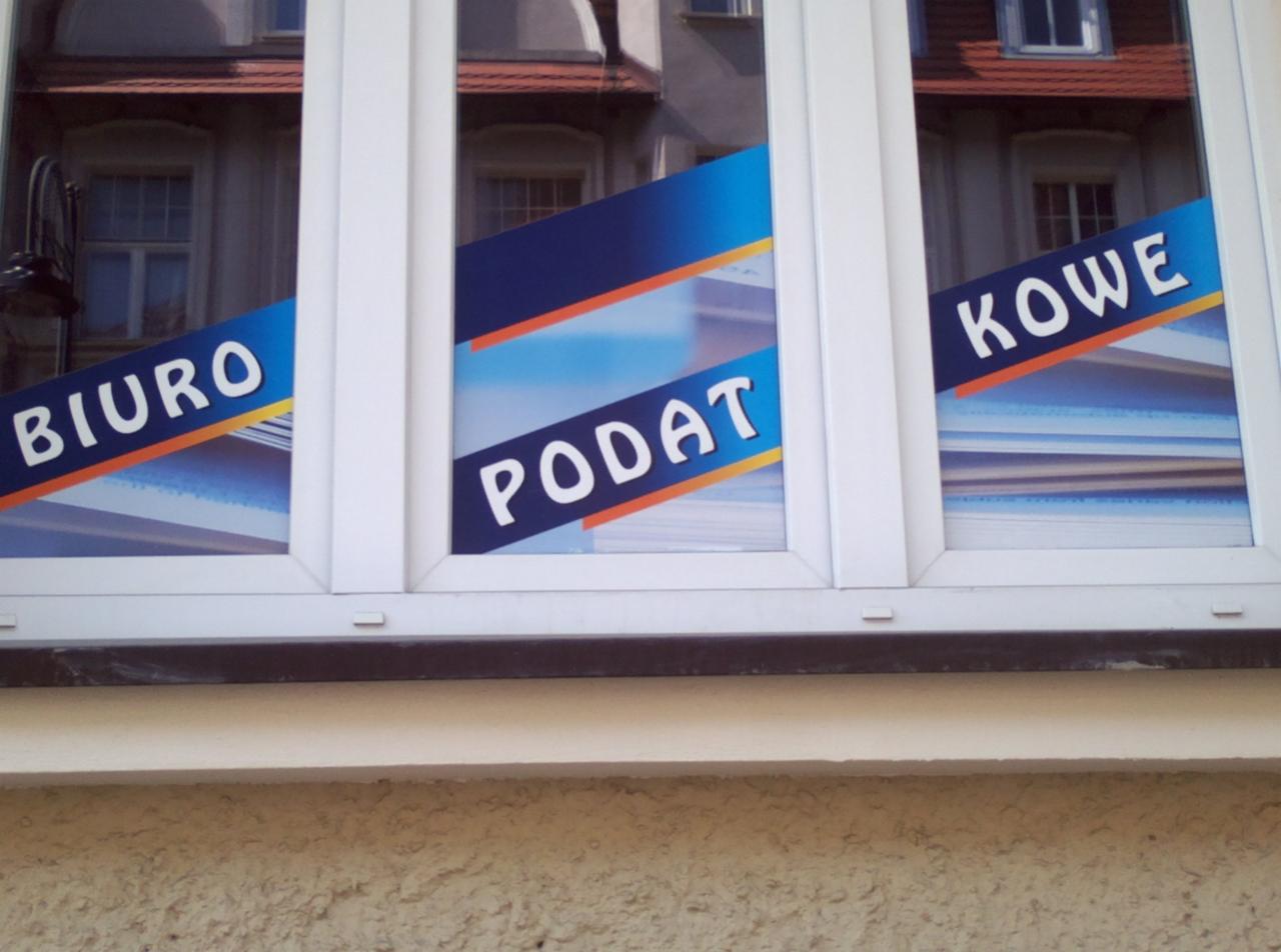 Okna biura rachunkowego z naklejonymi foliami z napisami: Biuro, Podatki, Kowe. W szybach odbija się budynek naprzeciwko.