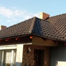 Brązowy dach z dachówki ceramicznej na domu jednorodzinnym z ceglanym kominem, widoczne rynny i obróbki blacharskie, w tle błękitne niebo.