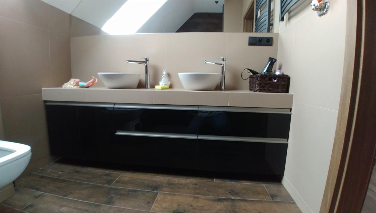 Łazienka z dwoma umywalkami nablatowymi na długim, czarnym blacie z szufladami, brązowe płytki podłogowe, beżowe ściany, zabawka leżąca obok umywalki.