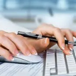 Dłonie kobiety piszącej na klawiaturze komputera, obok myszka i dokumenty, w tle monitor.