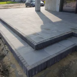 Nowo ułożony, szary chodnik z kostki brukowej z dwoma stopniami, obrzeża wykończone ciemniejszą kostką. W tle częściowo widoczny samoch&oacute;d i budynek.