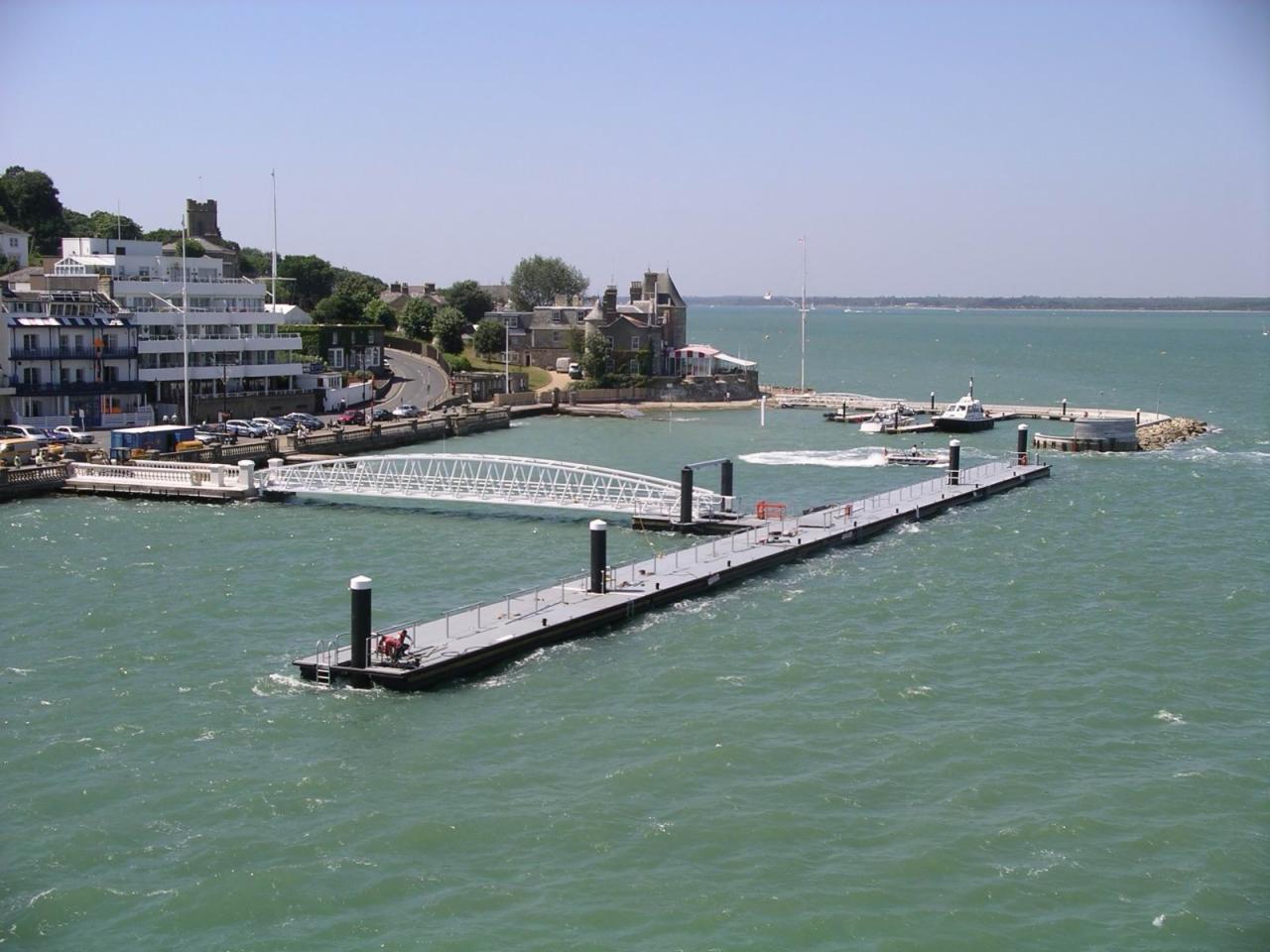 Długie, pływające molo z czarnymi słupkami w Warszawie, połączone z brzegiem białym mostem łukowym, widok z góry na turkusową wodę i zabudowania w tle.