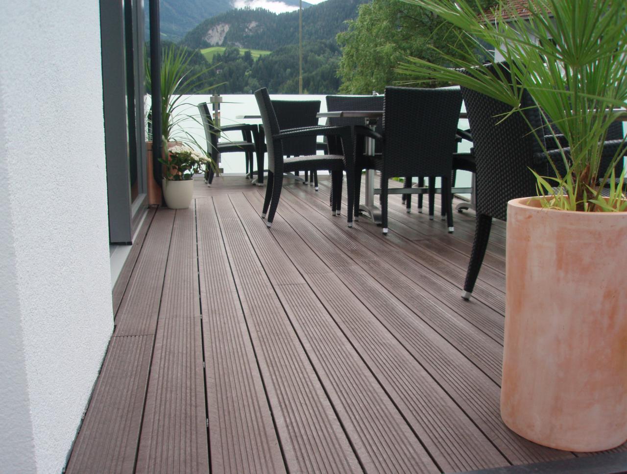 Kompozytowy taras w kolorze ciemnego drewna z widokiem na góry, wyposażony w czarne meble ogrodowe i rośliny w donicach z terakoty.