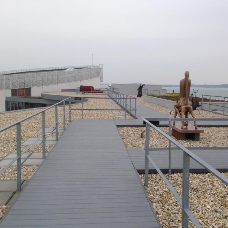 Szary pomost z desek kompozytowych na dachu budynku z żwirowym podłożem, metalowe barierki, rzeźby i widok na wodę w tle.