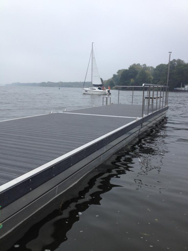 Pomost kompozytowy z metalową barierką na jeziorze, w tle żaglówka w pochmurny dzień.