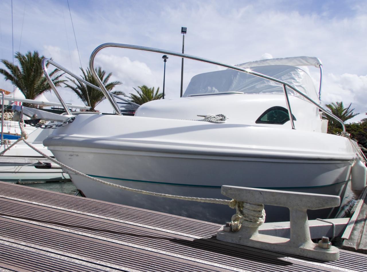 Biała łódź motorowa zacumowana przy drewnianym pomoście, liny cumownicze przymocowane do metalowej knagi, w tle palmy i błękitne niebo.