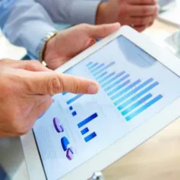 Mężczyzna wskazuje palcem na tablet z wyświetlonym wykresem słupkowym, filiżanka kawy i szklanka wody na biurku w tle.