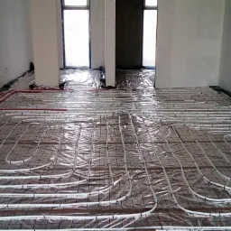 Rozłożona instalacja ogrzewania podłogowego na folii izolacyjnej, widoczne rury grzewcze w równych odstępach, przygotowane do zalania wylewką, w tle surowe ściany i okna.