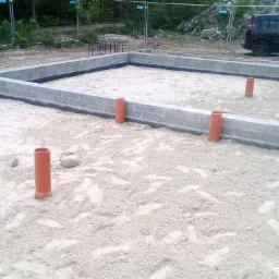 Fundamenty budynku w trakcie budowy, widoczne bloczki betonowe, izolacja przeciwwilgociowa oraz pionowe rury kanalizacyjne wystające z piasku.
