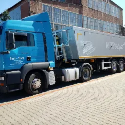 Niebieska ciężarówka MAN z naczepą MEGA Light na brukowanym placu, widoczny budynek przemysłowy w tle.