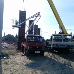 Montaż szalunków na budowie z użyciem podnośnika z platformą roboczą i dźwigu, widoczny czerwony bus z żółtymi kaskami w kabinie i numerem rejestracyjnym WPN UY71, w tle hałda ziemi i fragmenty...