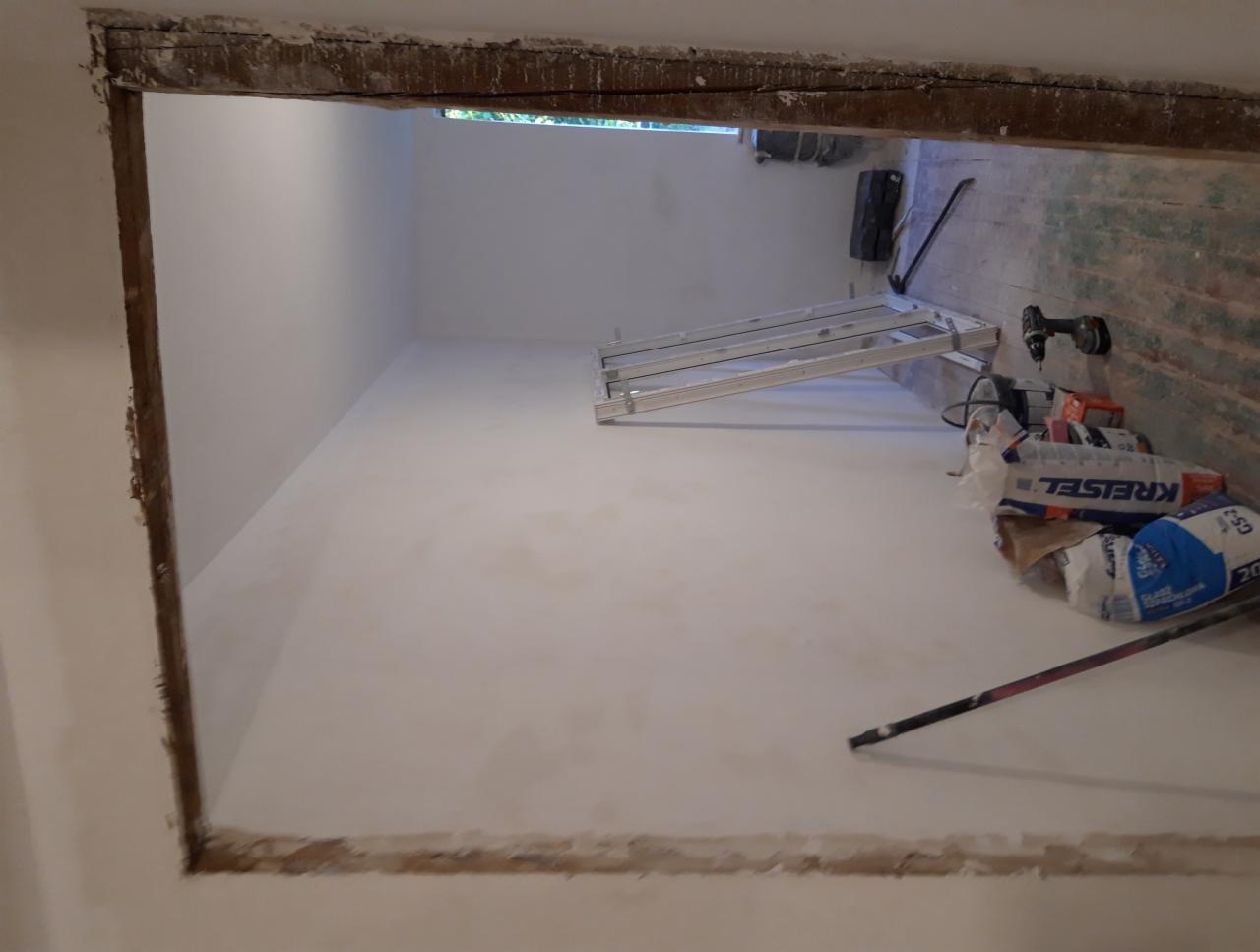 Przygotowana do malowania ściana w pomieszczeniu z widocznymi workami z zaprawą, drabiną, wiertarką i narzędziami malarskimi, ujęcie z perspektywy ościeżnicy.