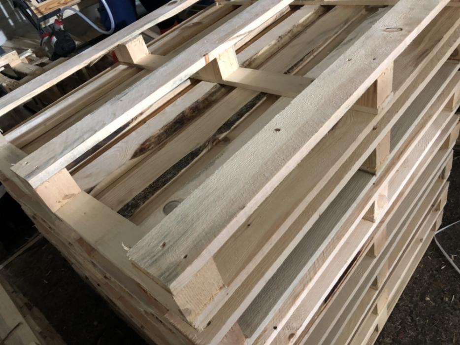 Stos jasnej, surowej tarcicy na paletach, widoczne ślady po korze na niektórych deskach.