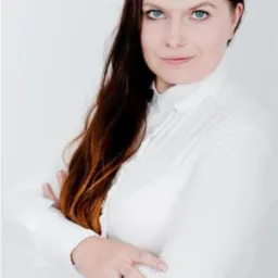 Portret kobiety o jasnych oczach, z długimi, brązowymi włosami i w białej koszuli, pozującej na jasnym tle z założonymi rękoma.