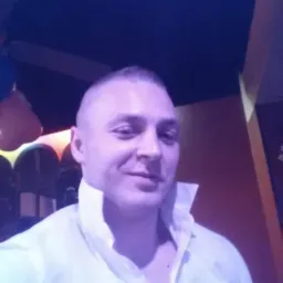 Portret mężczyzny w białej koszuli z rozpiętym kołnierzykiem, uśmiechniętego, na rozmytym tle wnętrza z dekoracjami.