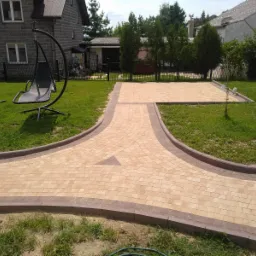 Ułożona ścieżka z kostki brukowej w kształcie litery Y, z obrzeżami w kolorze ciemnego brązu, prowadząca do tarasu z kostki, w tle dom z szarej cegły i wiszące krzesło.