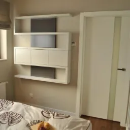 Nowoczesne, białe drzwi wewnętrzne z pionowym, matowym przeszkleniem w sypialni z beżowymi ścianami, drewnianą podłogą i zabudową meblową na ścianie.