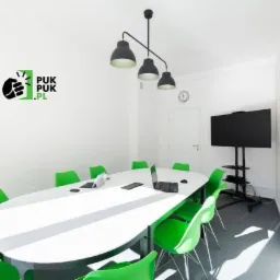Sala konferencyjna z białym stołem, zielonymi krzesłami, laptopami, tablicą suchościeralną i logo firmy na ścianie.