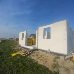 Wznoszenie ścian parterowego domu murowanego z bloczków Ytong na fundamencie, widoczne palety z bloczkami, w tle zielona trawa i błękitne niebo.