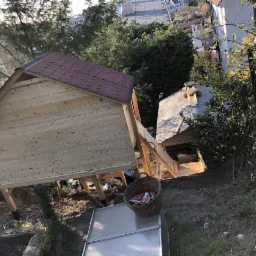Dwa domki drewniane na podwyższeniu w ogrodzie, jeden z czerwonym dachem, drugi z szarym, połączone drewnianą ścieżką, widok z góry na zbocze.