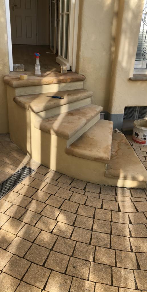 Wykończone schody wejściowe z beżowego kamienia, z widocznymi śladami po czyszczeniu, prowadzące do otwartego wejścia, z kostką brukową na podłożu i narzędziami pozostawionymi na stopniach.