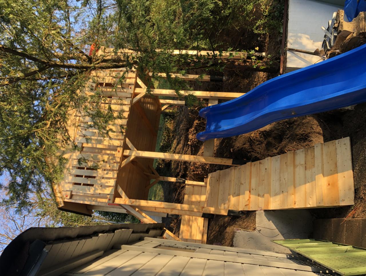 Drewniany plac zabaw dla dzieci w trakcie budowy: domek na drzewie na drewnianych podporach z niebieską zjeżdżalnią, widok z dołu na tle zieleni.