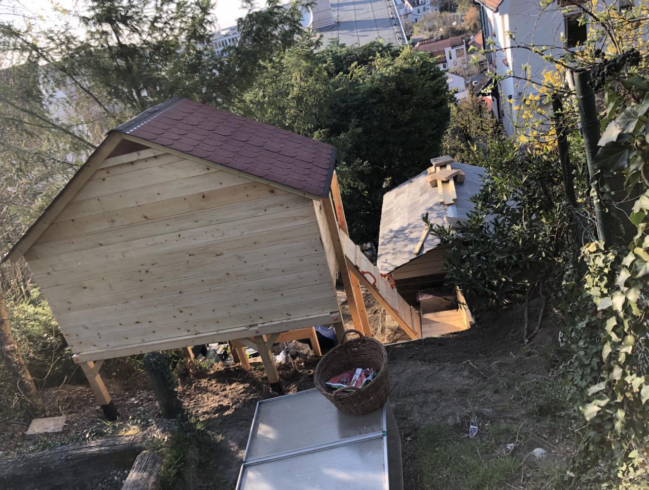 Dwa domki drewniane na podwyższeniu w ogrodzie, jeden z czerwonym dachem, drugi z szarym, połączone drewnianą ścieżką, widok z góry na zbocze.