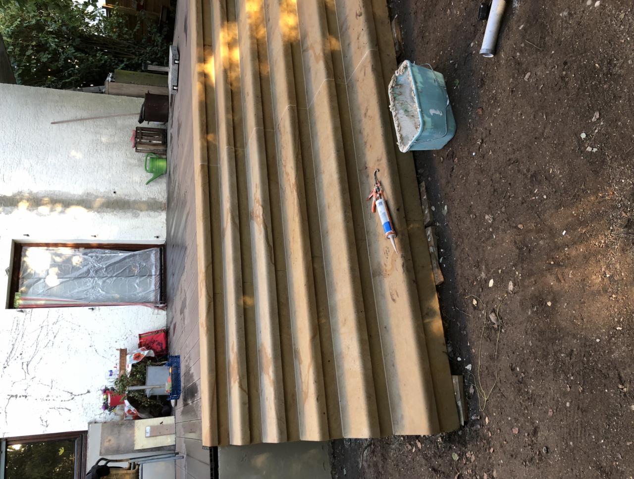 Fragment konstrukcji z profili, prawdopodobnie pergoli, z widocznymi narzędziami i pojemnikiem na brudnym podłożu, w tle budynek z oknem zabezpieczonym folią.