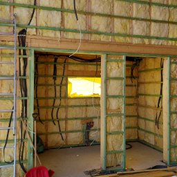 MK Projekt - Wnętrze budynku w trakcie izolacji: ściany z wełną mineralną, drewniana konstrukcja, instalacja elektryczna w peszlach, drabina aluminiowa oparta o ścianę, okno zabezpieczone folią.