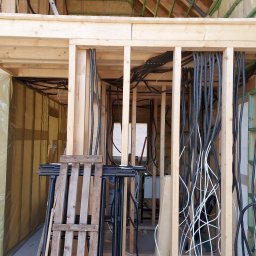 MK Projekt - Wnętrze w trakcie budowy z widoczną drewnianą konstrukcją szkieletową, licznymi przewodami elektrycznymi w peszlach w kolorze czarnym i białym, biegnącymi pionowo wzdłuż ścian, oraz elementami...