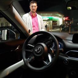 MK Projekt - Wnętrze samochodu Mazda CX-5 z widocznym kierowcą w koszuli w kolorze fuksji i jasnej marynarce, stojącego obok stacji benzynowej nocą.