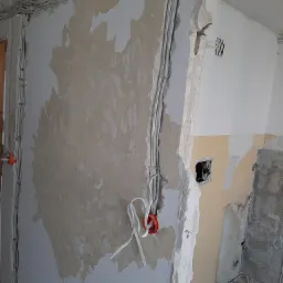 Instalacja elektryczna w trakcie remontu: widoczne kable prowadzone po ścianie, puszka elektryczna z wystającymi przewodami, fragment ściany z resztkami starej farby i tynku.