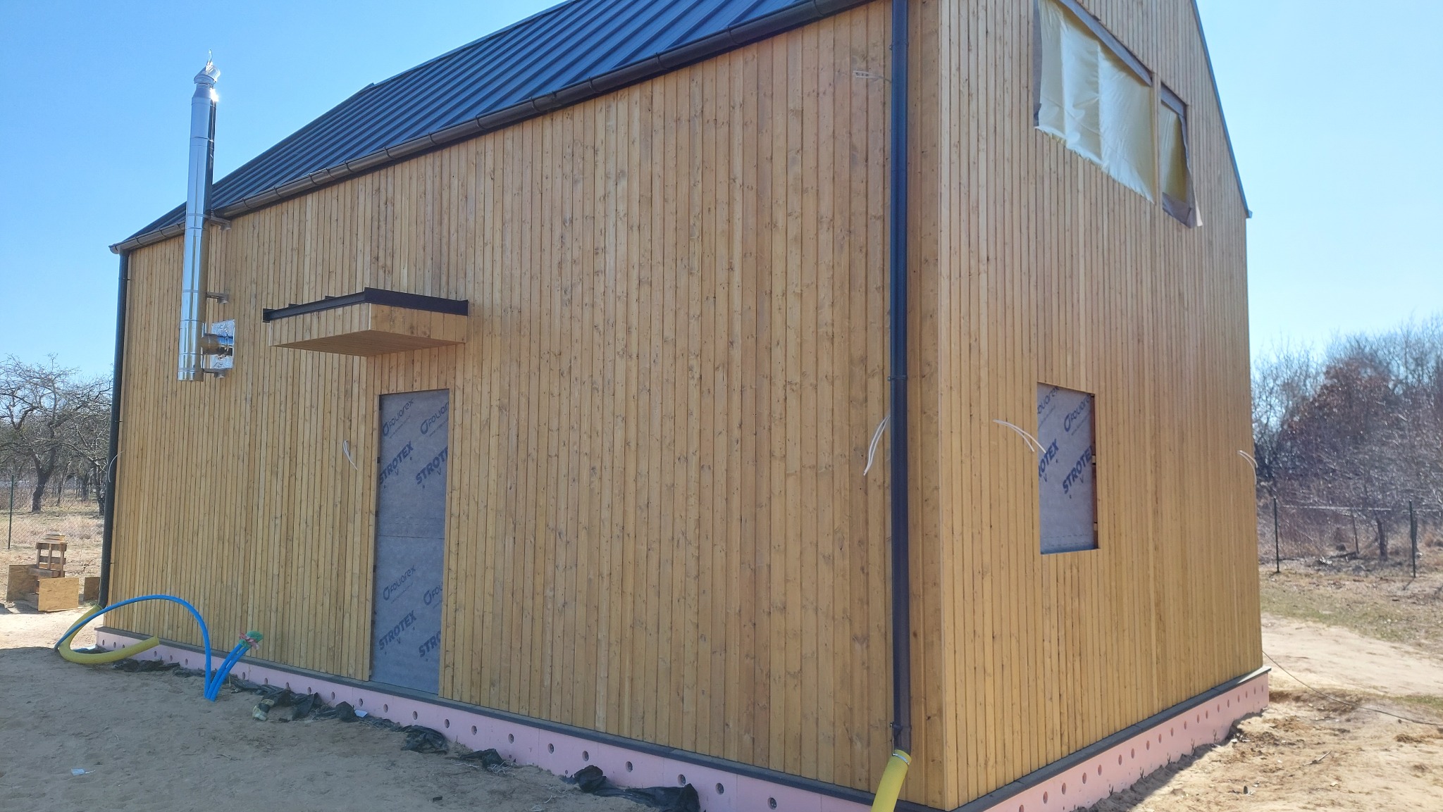 Surowy stan elewacji domu z drewnianą boazerią, widoczne okna i drzwi zabezpieczone folią, komin stalowy, rynna, ocieplenie fundamentów styropianem, teren budowy.