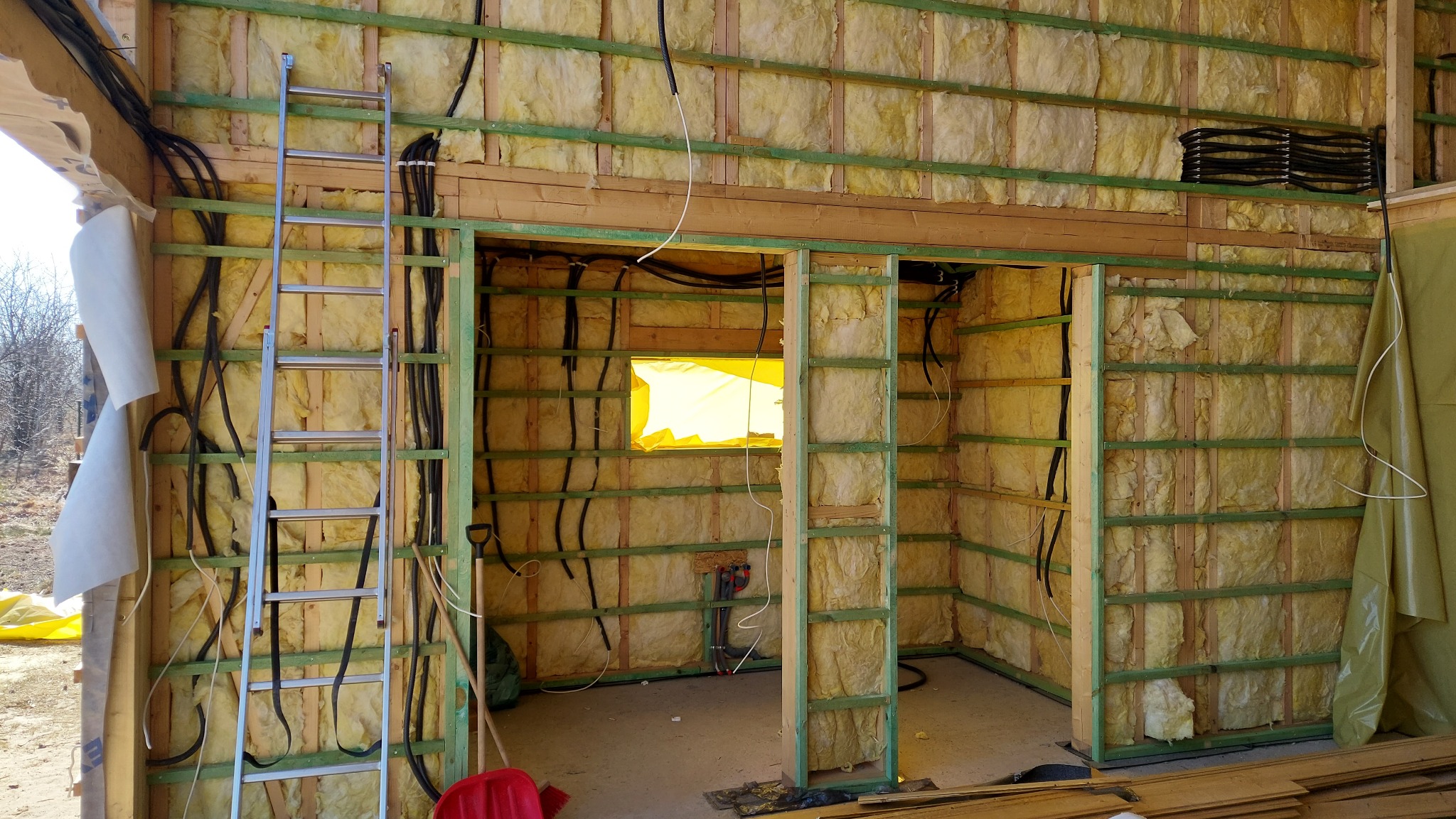 Wnętrze budynku w trakcie izolacji: ściany z wełną mineralną, drewniana konstrukcja, instalacja elektryczna w peszlach, drabina aluminiowa oparta o ścianę, okno zabezpieczone folią.