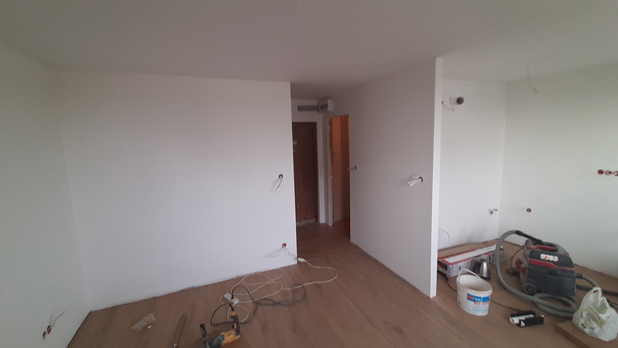 Wnętrze w trakcie remontu: świeżo pomalowane ściany na biało, widoczne kable elektryczne wystające ze ścian, odkurzacz przemysłowy, wiadro i narzędzia na podłodze z paneli.