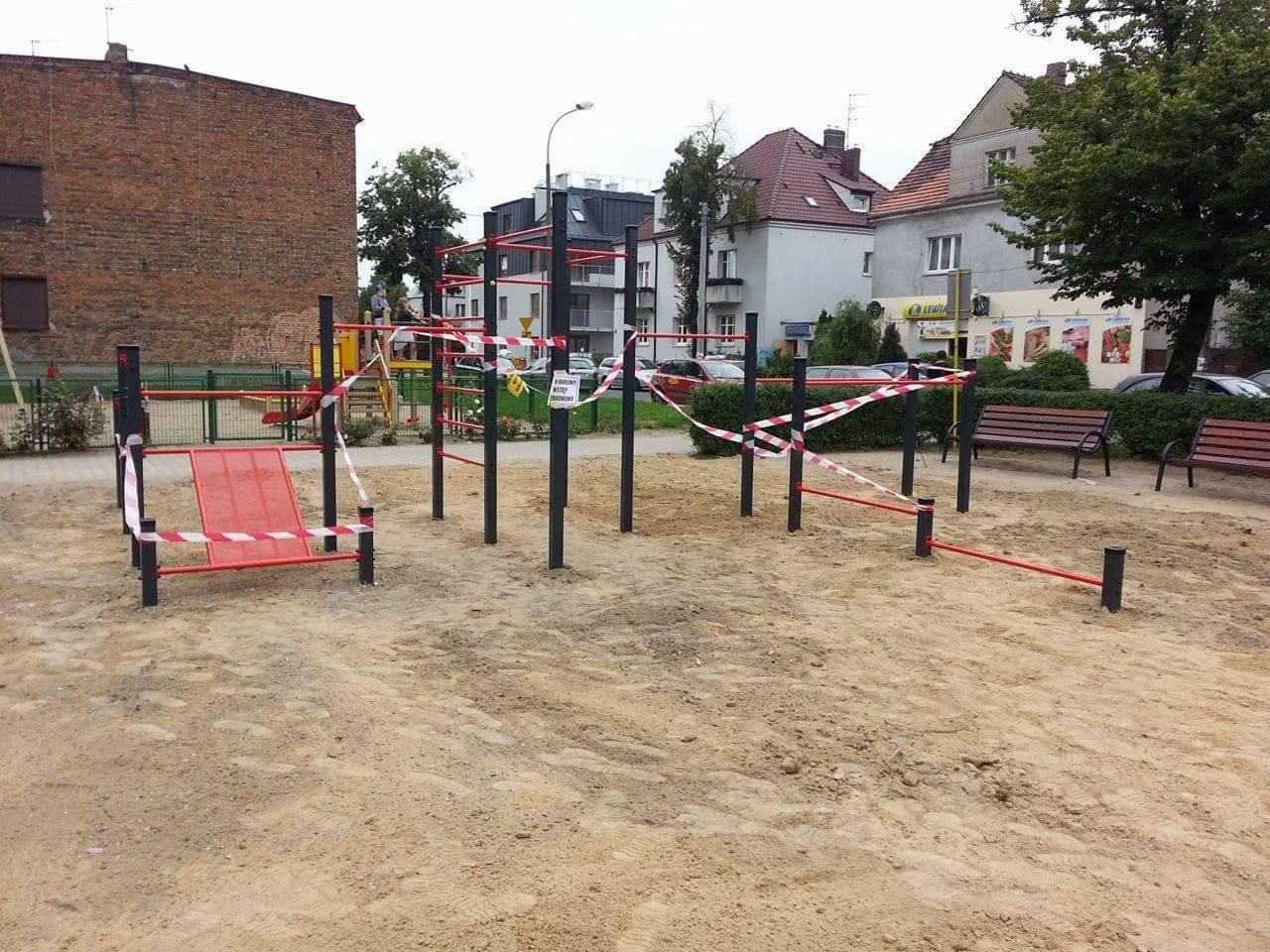 Nowo zainstalowany plac zabaw z metalowymi konstrukcjami do ćwiczeń, otoczony taśmą ostrzegawczą, na piaszczystym podłożu w otoczeniu budynków mieszkalnych i drzew.